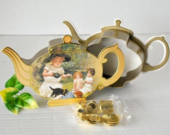 May include: A gold teapot with a picture of three children having tea in a garden. The teapot is open and the lid is off. There are gold-colored metal pieces in a plastic bag.