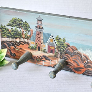 May include: A wooden key holder with a painted scene of a lighthouse on a rocky coastline. The holder has four hooks for hanging keys.