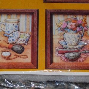May include: Two framed watercolor prints. The left print features a mug, soap, shaving brush, and pocket watch. The right print shows a vase of flowers, a bowl, and a hairbrush. The frames are brown.