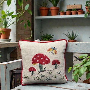 May include: A decorative pillow with a mushroom design. The pillow features a cream-colored background with red mushrooms and a butterfly. The pillow has a red border and is placed on a light blue bench.