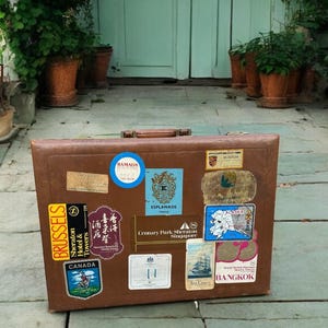 May include: A vintage brown suitcase covered in travel stickers, including labels for Brussels, Canada, and Bangkok. The suitcase is set on a stone patio in front of a light blue door and lush greenery.