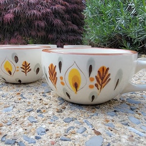 May include: Set of four white ceramic mugs with a brown, yellow, and gray floral design. The mugs have a rounded shape and a handle.