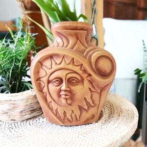 May include: A ceramic vase with a brown glaze features a raised design of a sun with a human face. The vase has a rounded shape and a wide opening.