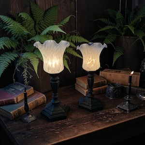 May include: Two electric lamps with black metal bases and frosted white glass shades featuring a ruffled edge. The lamps are positioned on a wooden surface alongside two small black metal candle holders and several vintage books.