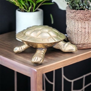 May include: A gold-colored metal turtle figurine with a detailed shell and legs. The turtle is sitting on a wooden table with a black background.