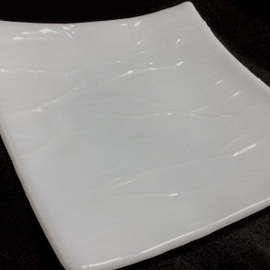 May include: A square, white ceramic plate with gently curved edges. The plate has a glossy finish and subtle, wave-like patterns across its surface. The design is simple and elegant, suitable for serving food.