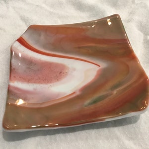 May include: A decorative, square-shaped glass dish with a unique, swirled pattern. The dish features a blend of colors, including white, shades of brown, and red. The edges are slightly curved, adding to its artistic appeal.