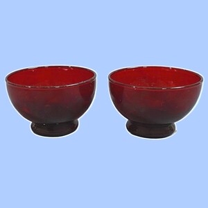 May include: Two matching deep red glass bowls with a smooth, rounded silhouette and a sturdy pedestal base. These translucent vessels feature a consistent crimson hue throughout the glass, showcasing a simple, functional design for serving food.
