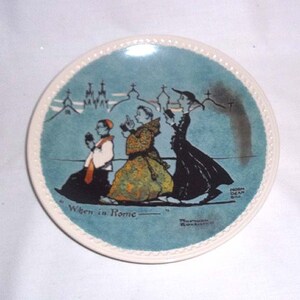 May include: A blue and white ceramic plate with a scene of four people walking in a city. The plate has a white border and the words "When in Rome" written on it.