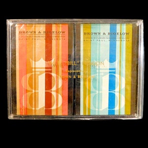 May include: A clear plastic case containing two decks of playing cards. The cards feature vertical stripes in orange, brown, and white on one deck, and blue, green, and white on the other. The text "BROWN & BIGELOW" is visible.