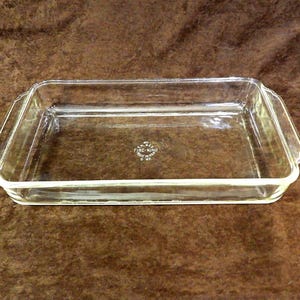 May include: A rectangular clear glass baking dish with integrated side handles. The center of the base features embossed text reading: 40, FIRE-KING, MADE IN U.S.A., 2 QT. The dish has a smooth, transparent finish and a simple, functional design.