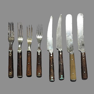 May include: A collection of vintage silverware, including forks and knives, arranged on a gray background. The forks have three or four tines and dark wooden handles. The knives have silver blades and wooden handles, some with decorative metal accents.