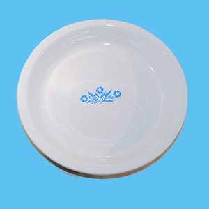 May include: White ceramic serving bowl with a light blue floral design in the center. The bowl has a wide rim and a shallow depth. The floral design features stylized flowers and leaves. The bowl is set against a light blue background.