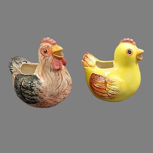 May include: Two ceramic chicken planters. One is a hen with a pink comb, wattle, and beak, and black and gray wings. The other is a yellow chick with orange and brown wing details and a pink comb. Both have openings for plants.