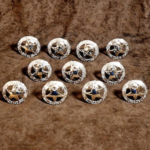 May include: A collection of eleven silver-colored buttons. Each button features a raised star design in the center, surrounded by a decorative border. The buttons are round and have a shiny, metallic finish, suitable for clothing or craft projects.