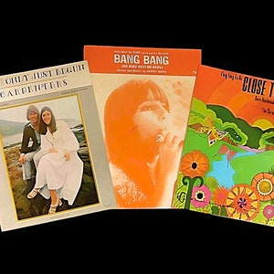 May include: Three sheet music covers. The first cover is light blue with a photo of a couple in front of a body of water. The title is "We've Only Just Begun" by The Carpenters. The second cover is orange with a photo of a woman with long dark hair. The title is "Bang Bang" by Nancy Sinatra. The third cover is green with a butterfly and flowers. The title is "Close To You" by The Carpenters.