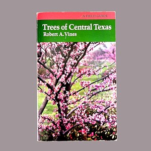 May include: A field guide titled "Trees of Central Texas" by Robert A. Vines. The cover features a photograph of a tree with pink blossoms against a green and brown background. The top of the book has a red stripe.