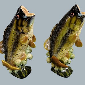 May include: Two ceramic fish sculptures with open mouths, featuring a color palette of black, yellow, and brown. The fish are posed as if leaping from water, with detailed fins and scales. The base is a mix of green and brown.