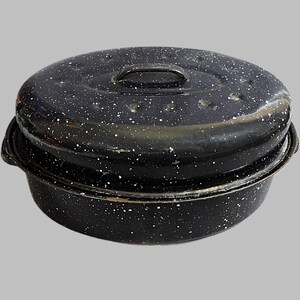 May include: An oval-shaped black roasting pan with a speckled white enamel finish. The pan features a matching lid with a central handle and integrated side handles on the base, showcasing a classic design with visible wear along the edges.