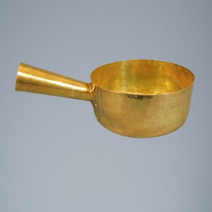 May include: A gold-colored ladle with a rounded bowl and a long, tapered handle. The ladle appears to be made of metal and has a shiny, reflective surface. The handle is attached to the bowl with rivets. The background is a solid, neutral color.