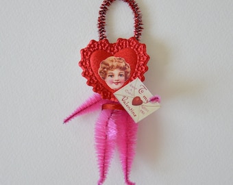 May include: A Valentine's Day ornament in the shape of a heart with a vintage image of a child's face. The heart is red with a decorative edge and a red pipe cleaner loop. Pink pipe cleaners form the arms and legs, and it holds a small envelope.