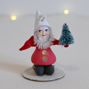 Puede incluir: Una figurita de elfo navideño vintage con un sombrero blanco y un abrigo rojo, sosteniendo un pequeño árbol de pino verde. El elfo está sentado sobre una base blanca.