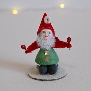 May include: A vintage Christmas gnome figurine with a red hat, white beard, and green pants. The gnome is holding two red jingle bells in its hands and is sitting on a glittery base.
