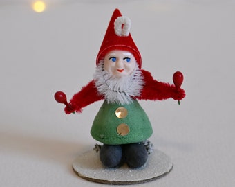 May include: A vintage Christmas gnome figurine with a red hat, white beard, and green pants. The gnome is holding two red jingle bells in its hands and is sitting on a glittery base.