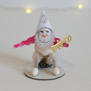 May include: A white and pink pipe cleaner gnome wearing a white hat with a gold trumpet. The gnome is sitting on a white glitter base.