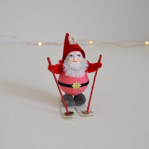 May include: A red and white gnome figurine on skis. The gnome is wearing a red hat with a white pom-pom and a white beard. The skis are red and white.