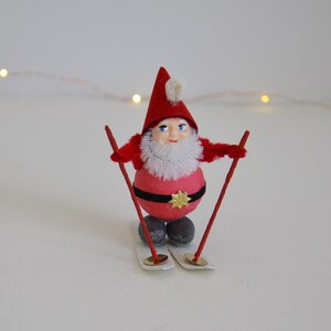 May include: A red and white cotton gnome wearing a red hat with a white pom-pom, holding red and white ski poles, and standing on white skis.