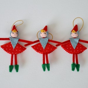 Op de afbeelding: Drie rode en witte kerstversieringen van vilt in de vorm van elven met pijpenragerarmen en -benen. Elke elf heeft een grijs vest van vilt, een geel haarstuk van vilt en een rode viltmuts.