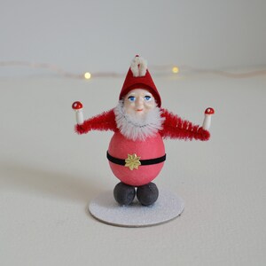 Könnte beinhalten: Eine rote und weiße Weihnachts-Gnom-Figur mit weißem Bart und roter Mütze. Der Gnom hält zwei rote und weiße Pilze in seinen Händen. Der Gnom steht auf einem weißen Sockel.