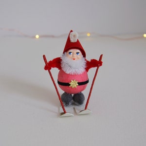 May include: A red and white Christmas gnome wearing a red hat with a white pom-pom and a white beard. The gnome is holding red and white ski poles and is standing on white skis.