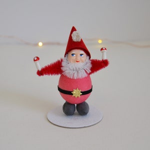 May include: A red and white Christmas gnome figurine with a white beard and a red hat. The gnome is holding two red and white mushrooms in its hands. The gnome is standing on a white base.
