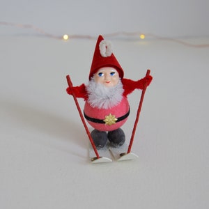 May include: A red and white Christmas gnome figurine wearing a red hat with a white pom-pom and holding red ski poles. The gnome is wearing a black belt with a gold star and is standing on white skis.