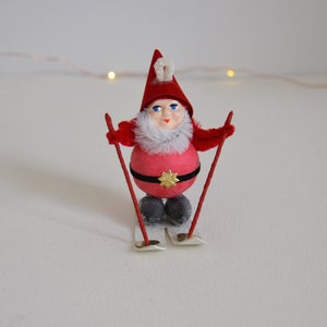 May include: A red and white Christmas ornament of a gnome wearing a red hat and holding ski poles. The gnome is standing on a pair of skis.