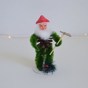 May include: A green and white Christmas gnome figurine with a red hat and a pine cone body. The gnome is holding a small broom.