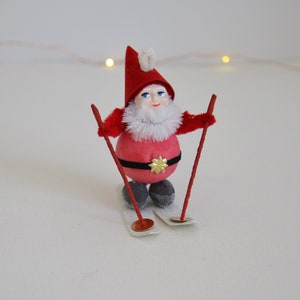 May include: A red and white felt Christmas gnome wearing a black belt with a gold star. The gnome is holding two red and white ski poles and is standing on a pair of white skis.