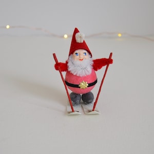 May include: A red and white Christmas gnome wearing a red hat with white trim and a white beard. The gnome is holding two red ski poles and is standing on a pair of white skis.