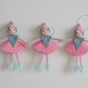 Op de afbeelding: Drie roze en grijze vilten ornamenten in de vorm van feeën met witte stippen, blauwe pijpenrager benen en zilveren draad halo's.