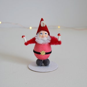May include: A red and white Christmas gnome figurine with a white beard and a red hat. The gnome is holding two red and white mushrooms in its hands. The gnome is standing on a white base.
