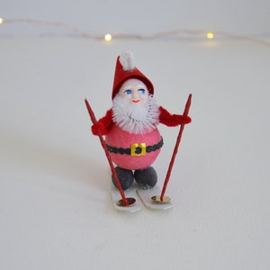 May include: A red and white Christmas gnome wearing a red hat and a black belt is skiing down a white slope. The gnome has a white beard and blue eyes.