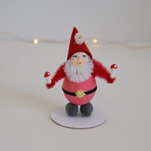 May include: A red and white Christmas gnome ornament with a white beard and a red hat. The gnome is holding two red and white mushrooms in its hands. The gnome is standing on a white base.