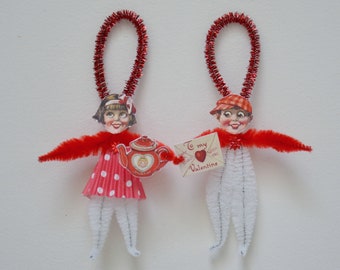 May include: Two Valentine's Day ornaments made from pipe cleaners and paper. The girl ornament is wearing a red and white polka dot dress and holding a red teapot. The boy ornament is wearing a red and white checkered hat and holding a card that says "To my Valentine".