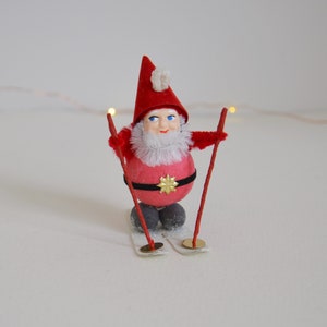 May include: A red and white Christmas gnome wearing a red hat with white trim and a white beard. The gnome is holding two red ski poles and is standing on a pair of white skis.