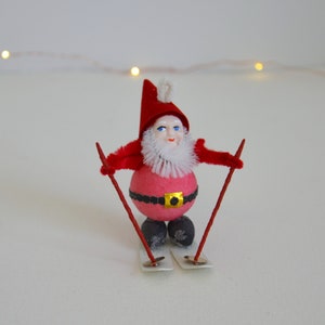 May include: A vintage Christmas ornament depicting Santa Claus skiing. The figure has a red hat and jacket, a pink body, and a white beard. The skis and poles are red. The ornament has a gold belt buckle and black feet.