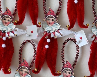 May include: A set of six red and white felt Christmas ornaments shaped like jesters. Each ornament has a white felt face with a red hat and a playing card attached to it. The playing cards are all aces with a red heart.