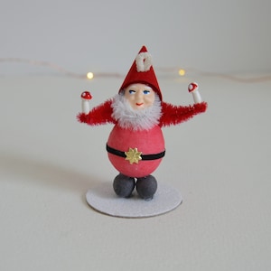 May include: A whimsical Christmas gnome figurine with a red body, hat, and arms, and a white beard and collar. The gnome holds mushroom-shaped ornaments. A black belt with a gold star buckle accents the waist. The figurine stands on a white, glittery base.