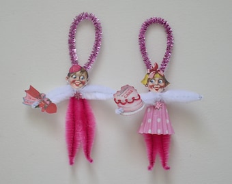 May include: Two pink and white pipe cleaner Valentine's Day ornaments. One ornament is a boy holding a Valentine's Day card that says "Be My Valentine". The other ornament is a girl holding a pink cake that says "Happy Valentine's Day Cake".